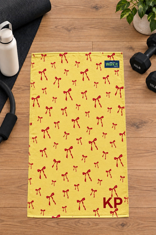 Yellow and red bow personalized gym towel with custom 'KP' initials, laid flat on a wood floor next to fitness gear.