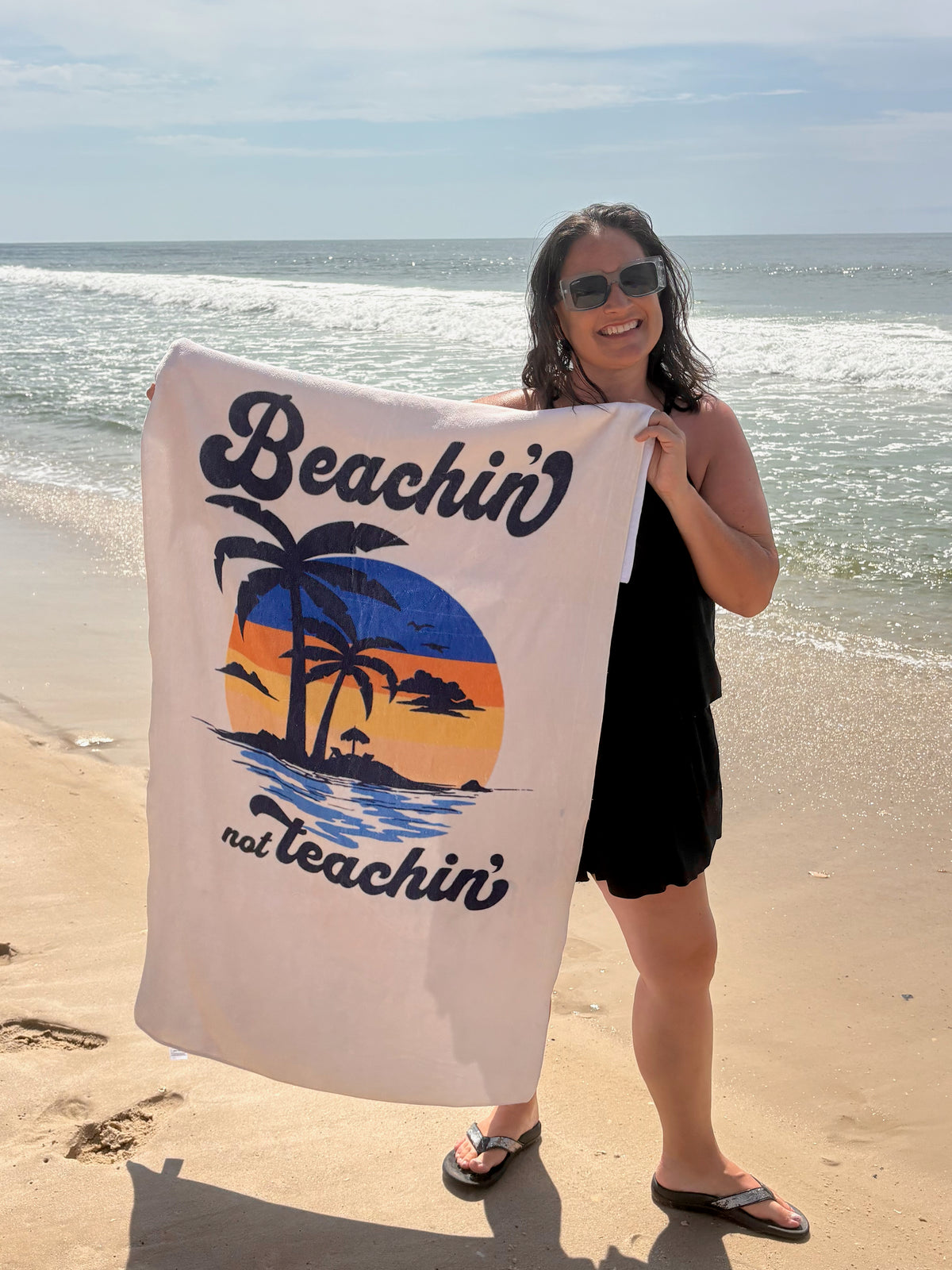 our 'beachin not teachin' sunset design beach towel being held by a young woman on a beach 