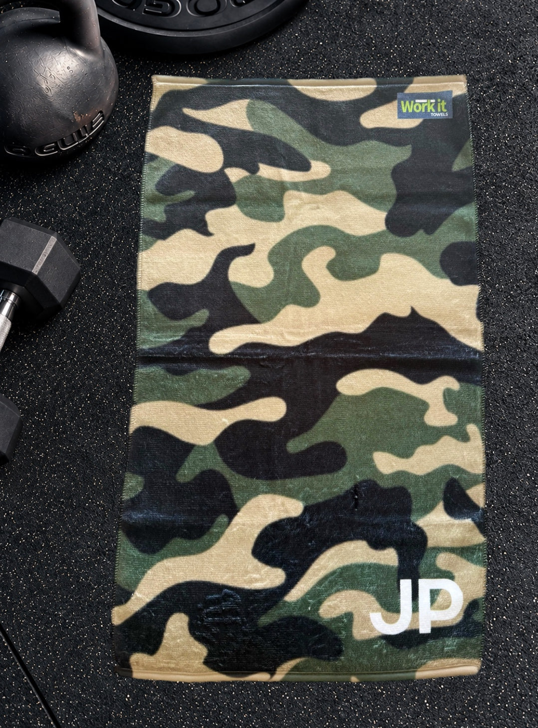 Green camouflage personalized gym towel with custom 'JP' initials, laid flat on a black gym floor with a kettlebell and dumbbell.