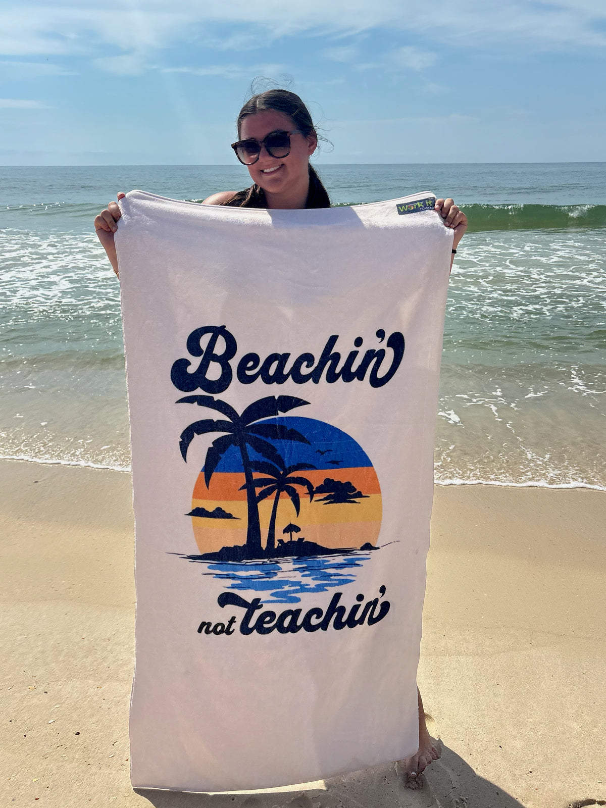 our 'beachin not teachin' sunset design beach towel being held by a young woman on a beach 