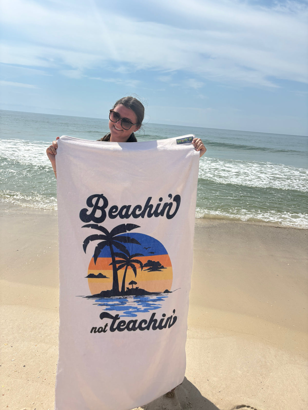 our 'beachin not teachin' sunset design beach towel being held by a young woman on a beach 