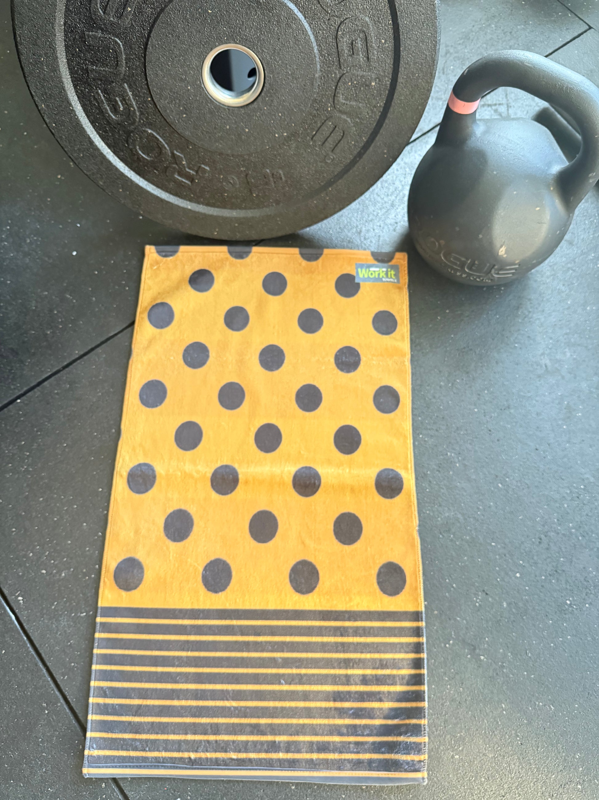 Yellow towel with black polka dots and stripes, placed on a tiled floor with a barbell and kettlebell in the background.