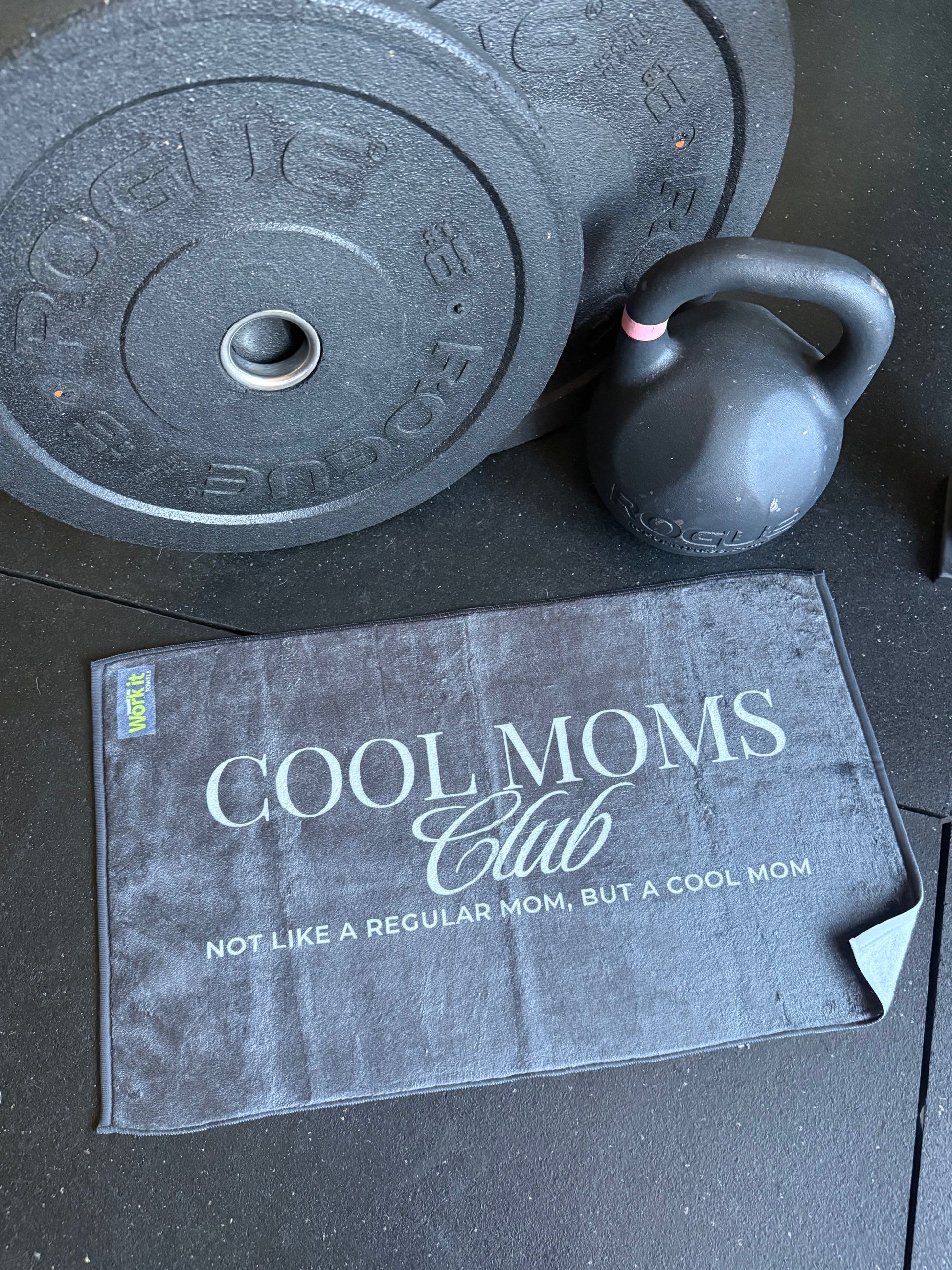 Kettlebell and weight plates on a surface with a 'Cool Moms Club' towel.