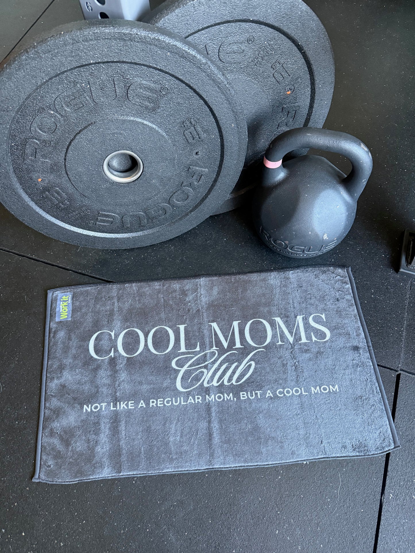 Kettlebell and weight plates on a surface with a 'Cool Moms Club' towel.