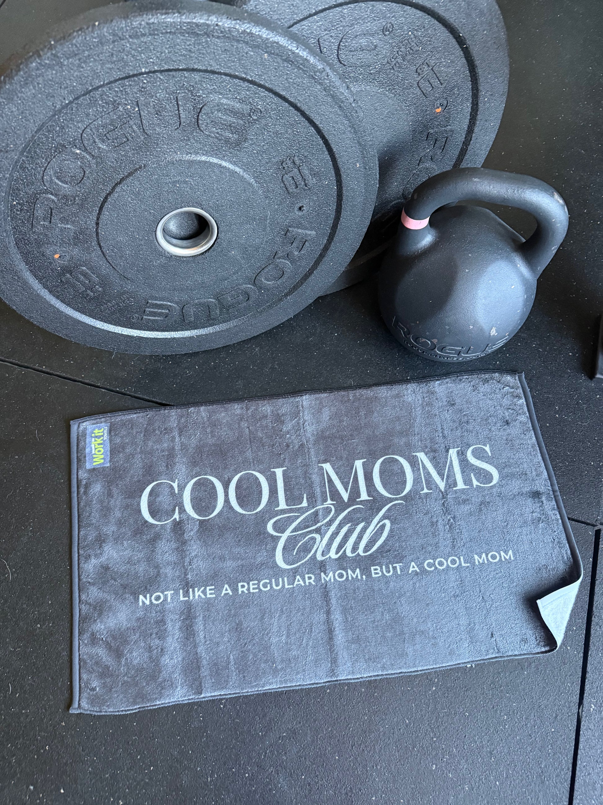 Black weight plate, kettlebell, and 'Cool Moms Club' towel on a black surface