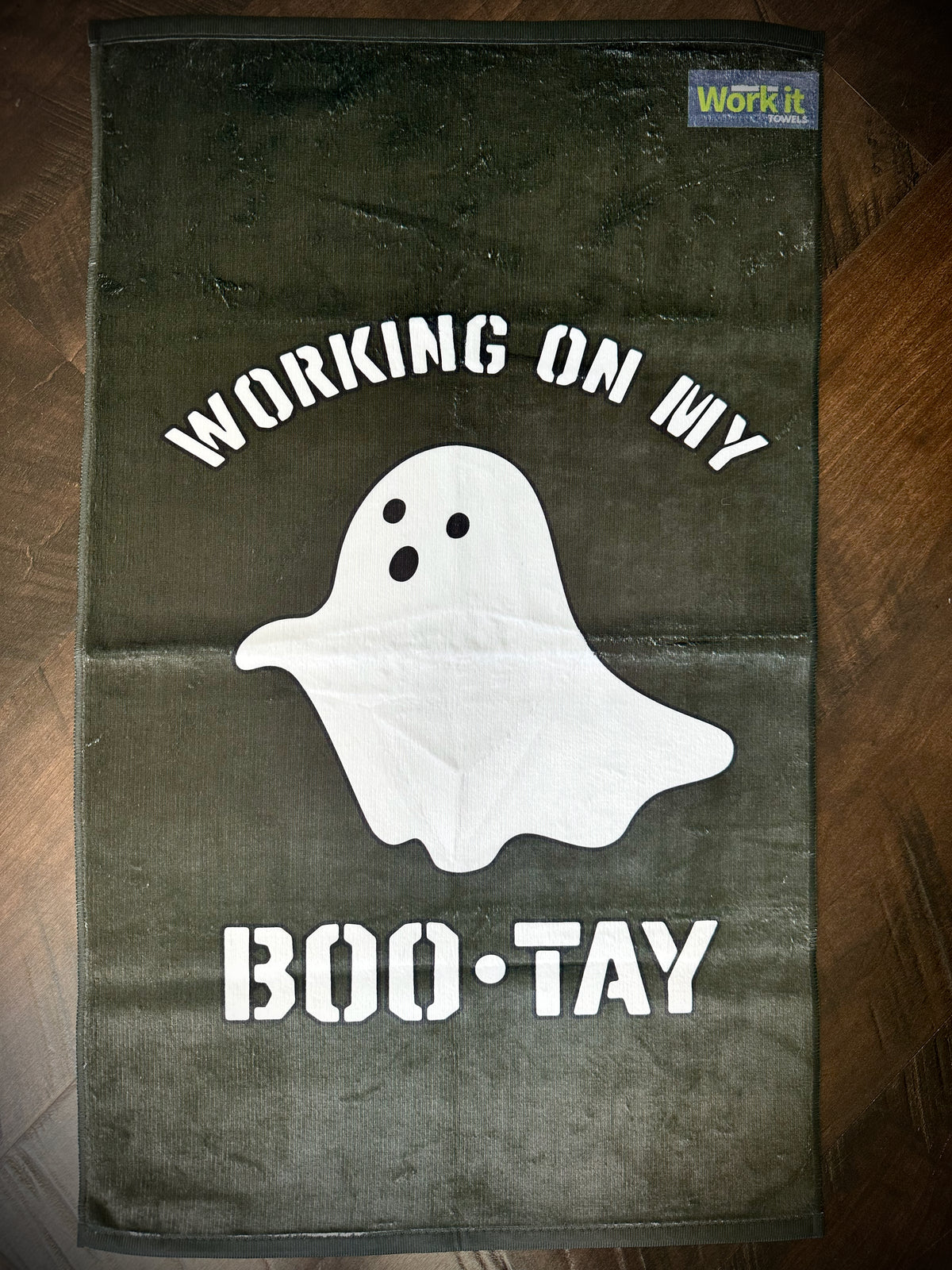Towel with ghost graphic and text 'Working on my boo-tay' on a wooden surface