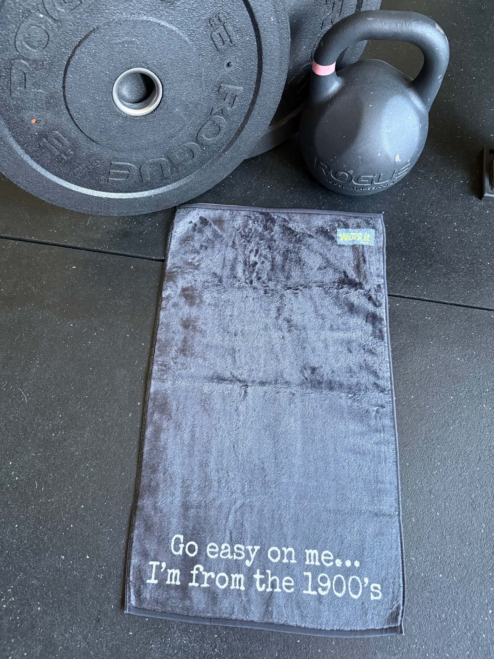 Black gym towel with humorous text on a gym floor with weights