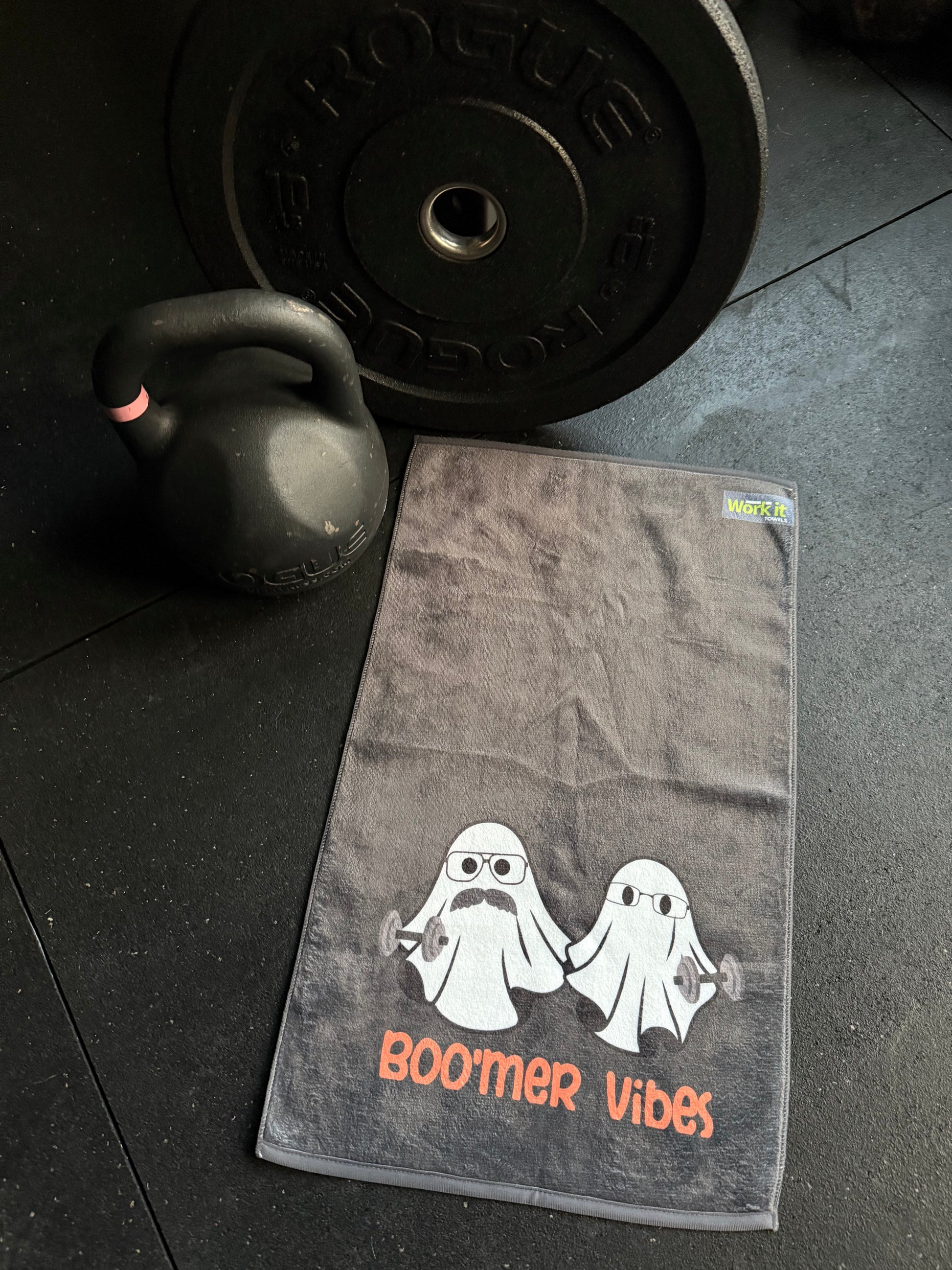 Gray towel with 'Boomer Vibes' logo on a gym floor with kettlebell and weight plate.