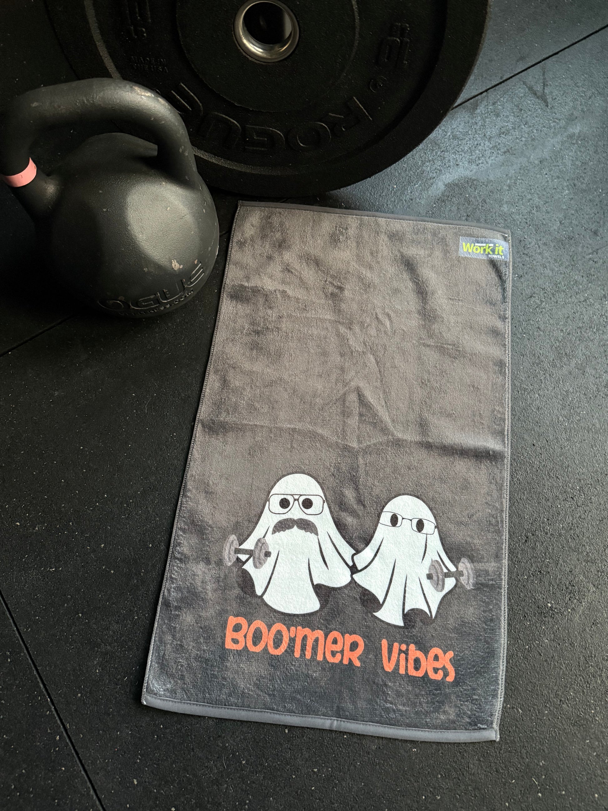 Gray towel with 'Boomer Vibes' logo on a gym floor with a kettlebell and weight plate.
