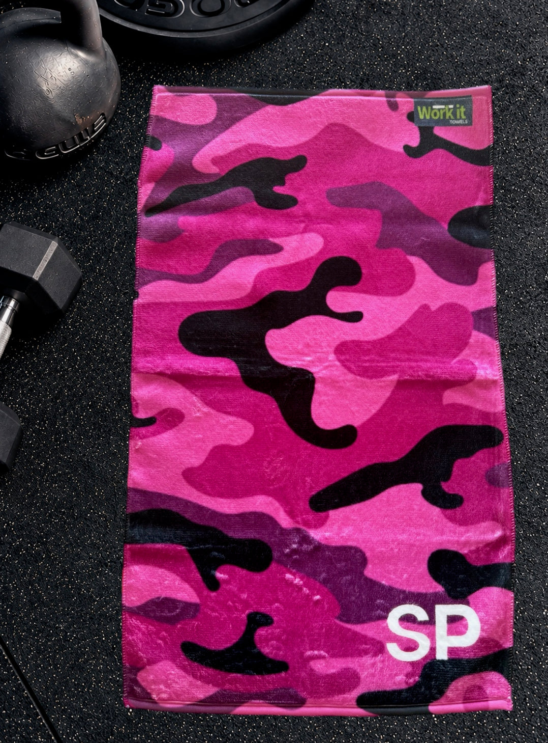 Pink camouflage personalized gym towel with custom 'SP' initials, laid flat on a black gym floor with dumbbells.