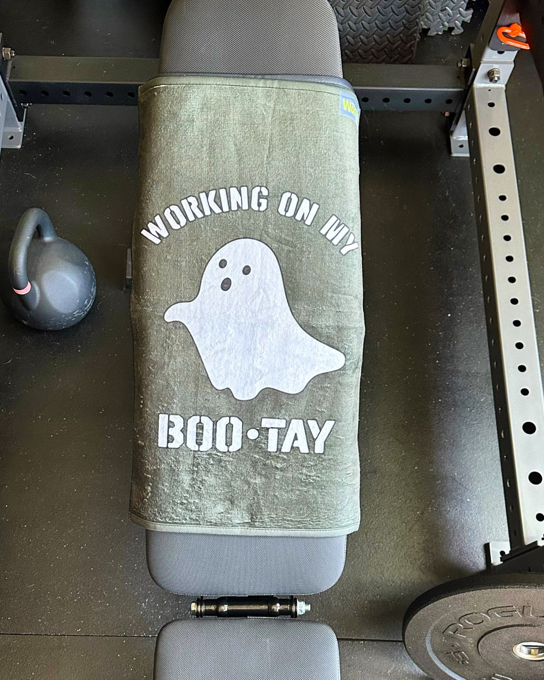 Gym bench with a towel featuring a ghost graphic and text on top, surrounded by gym equipment.