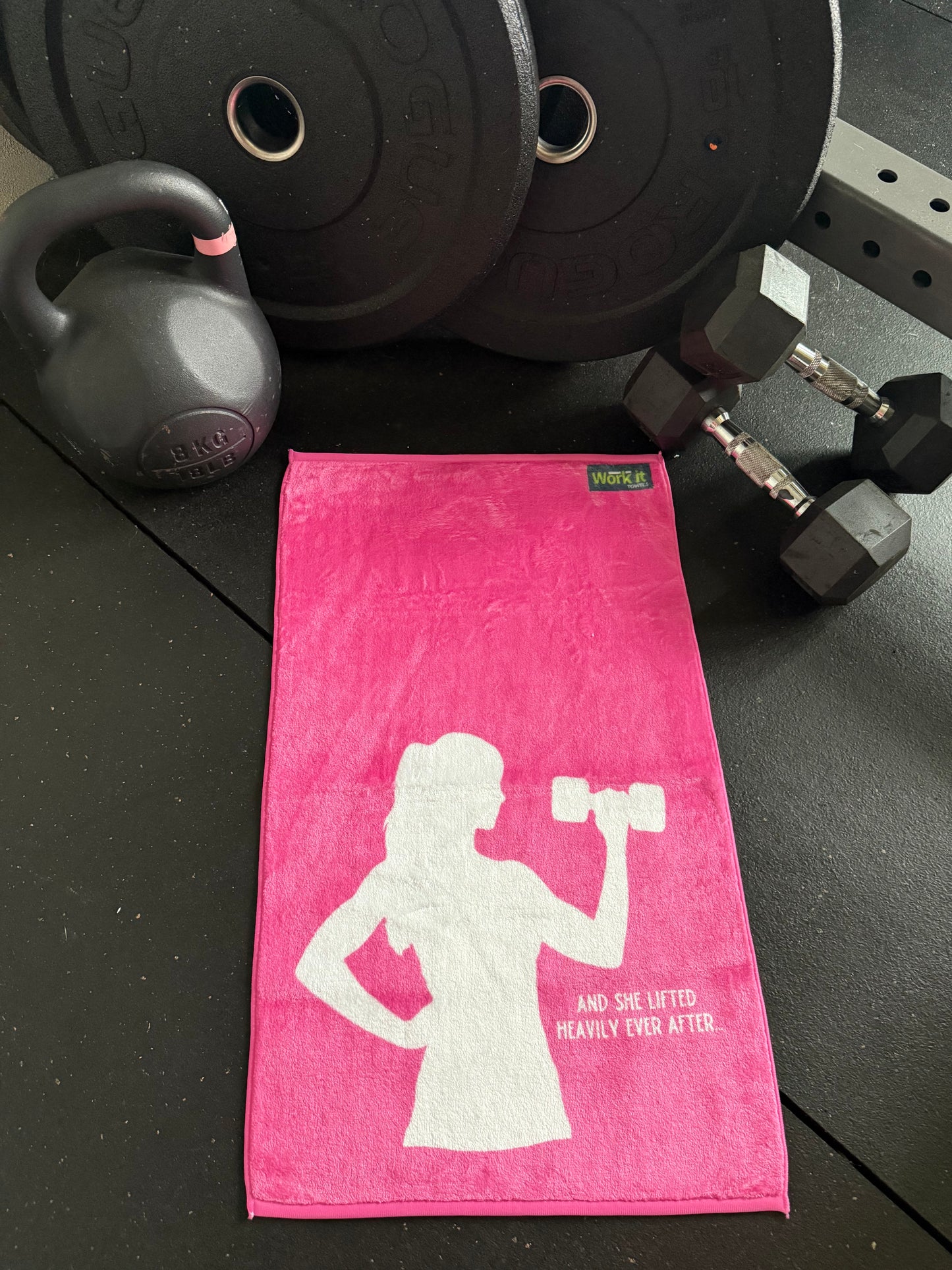 Pink towel with a white silhouette of a person lifting weights, placed on a black surface with dumbbells and weight plates.