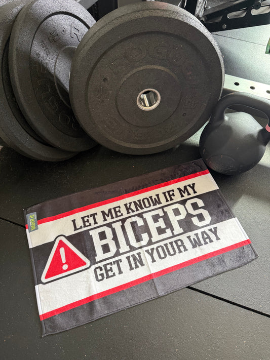 Stack of weight plates with a towel labeled 'Let me Know if my Biceps get in your way' on a gym floor.