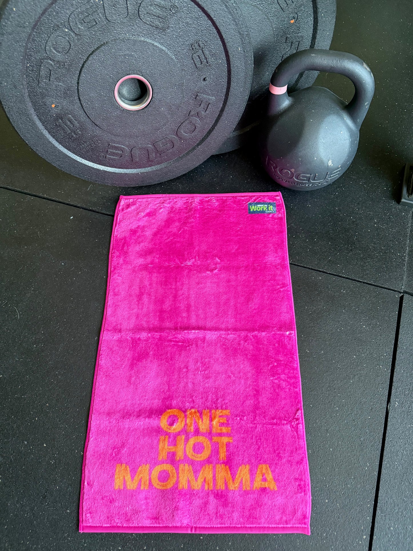Pink towel with 'One Hot Momma' text on a black floor with gym equipment.