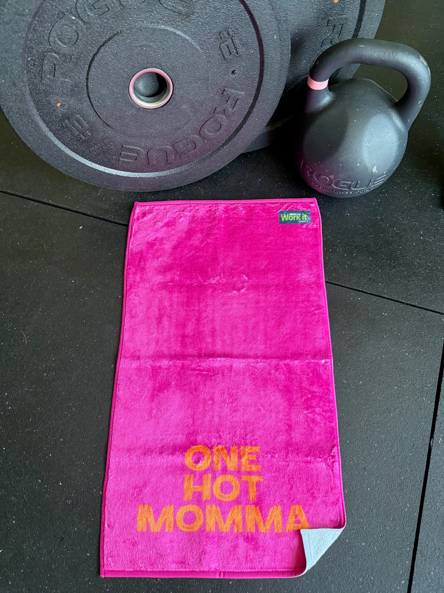 Pink towel with 'One Hot Momma' text on a black floor with gym equipment.