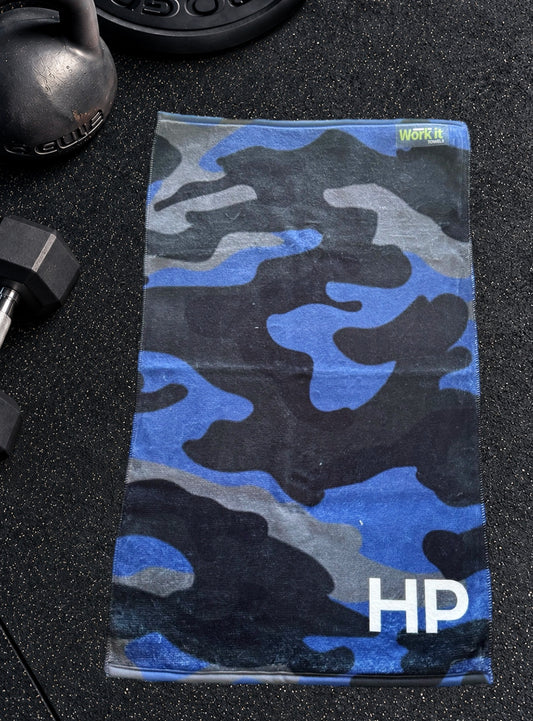 Blue camouflage personalized gym towel with custom 'HP' initials, laid flat on a black gym floor with a kettlebell and dumbbell.