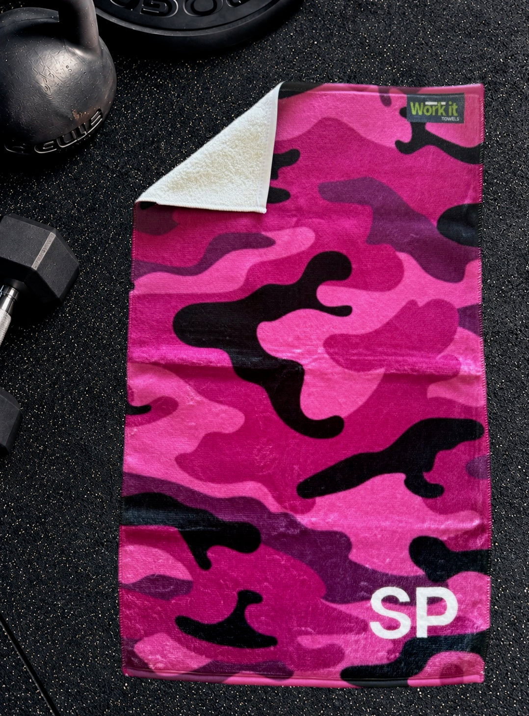 Pink camouflage personalized gym towel with custom 'SP' initials, one corner folded back to show the soft white terry reverse side.