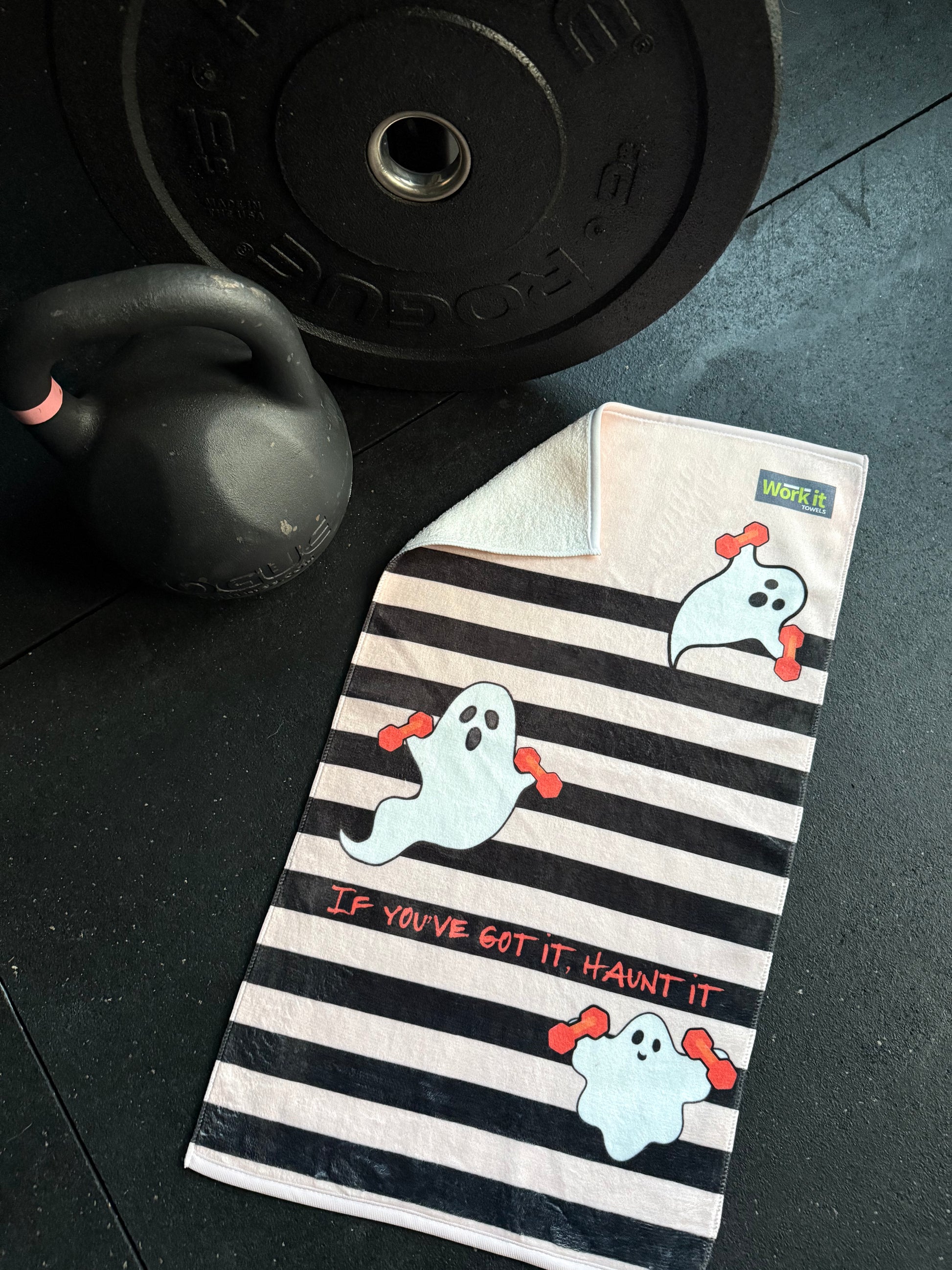 Cute gym towel with ghost design next to a barbell and kettlebell on a dark surface