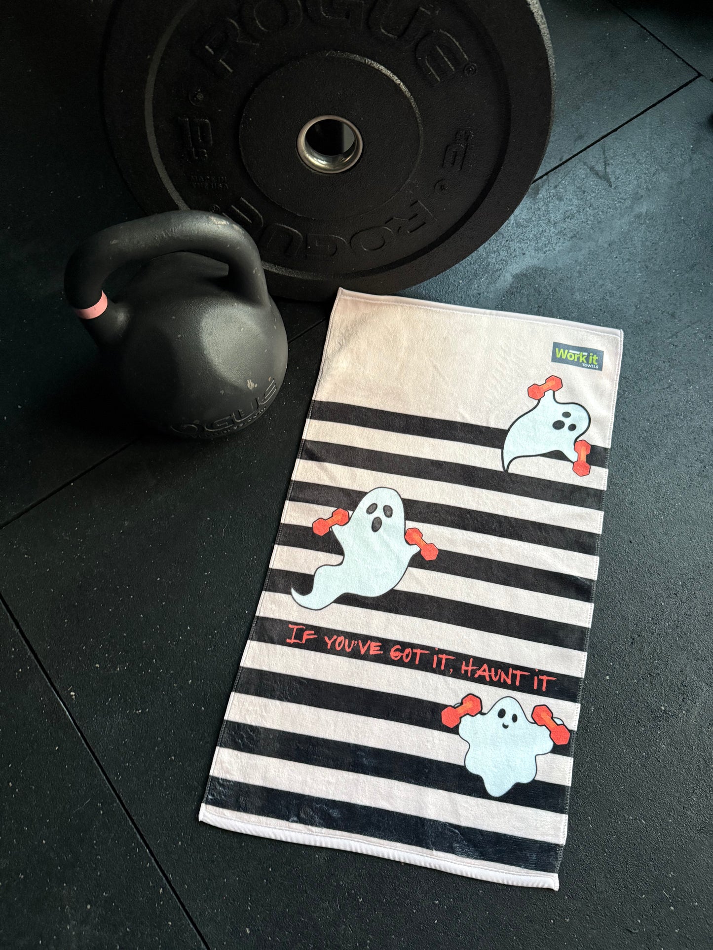 Striped pink towel with ghost design next to a kettlebell and weight plate on a dark surface