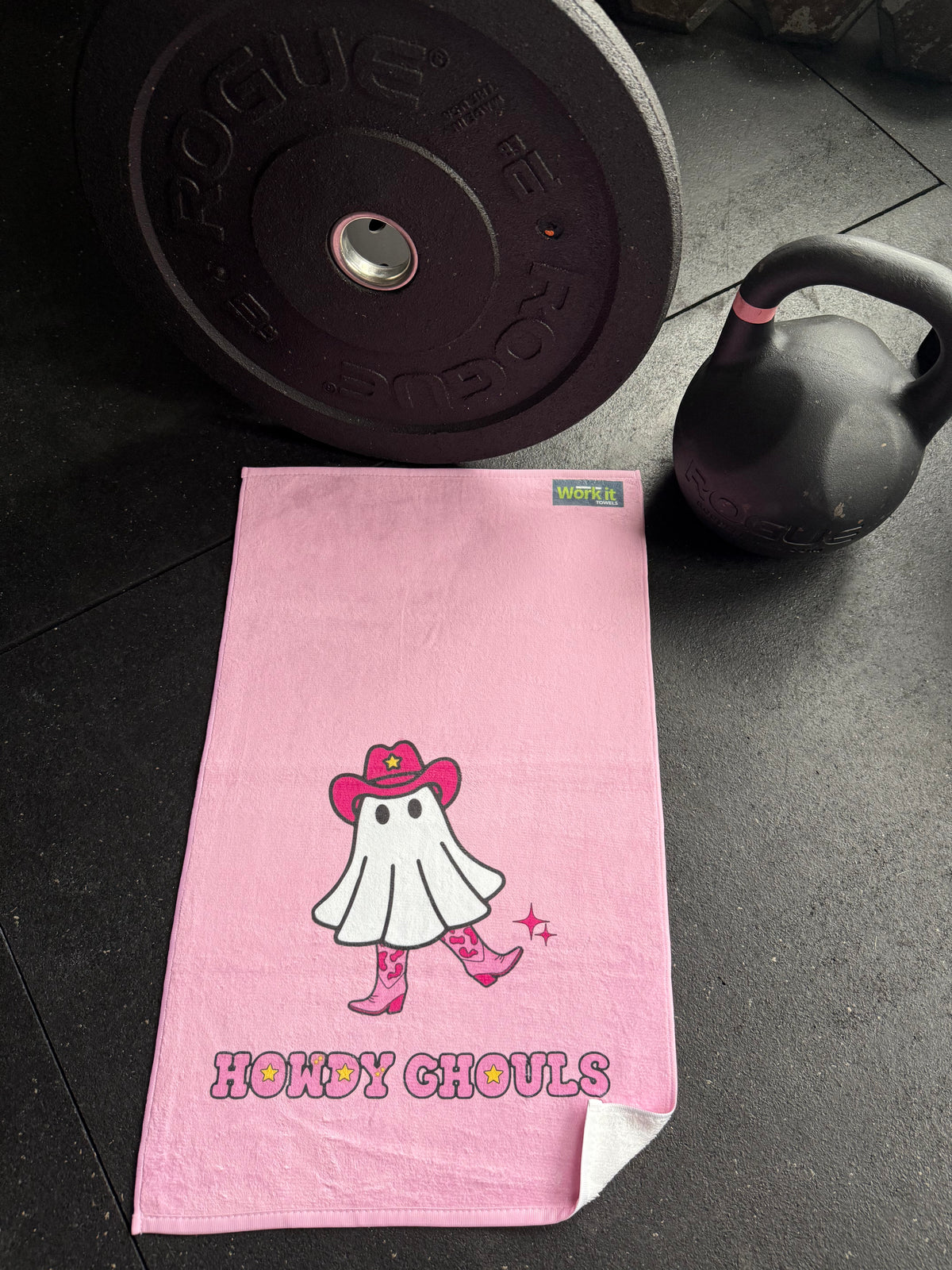 Pink towel with cartoon cowgirl  character and text on a gym floor with kettlebell and barbell.