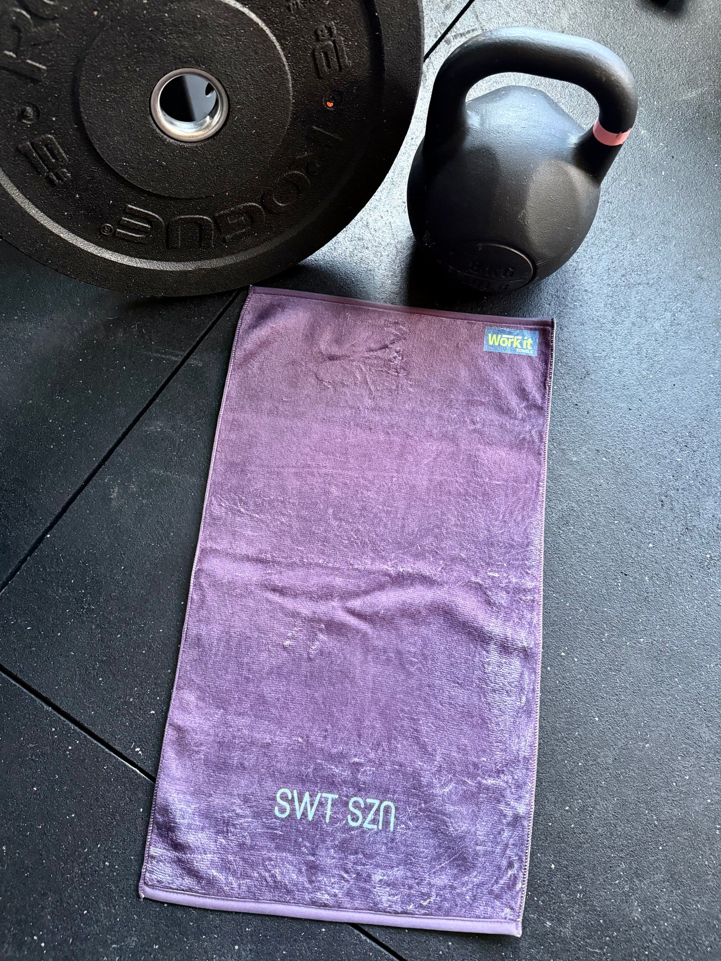 Purple towel with 'SWT SZN' branding on a black surface with gym equipment.