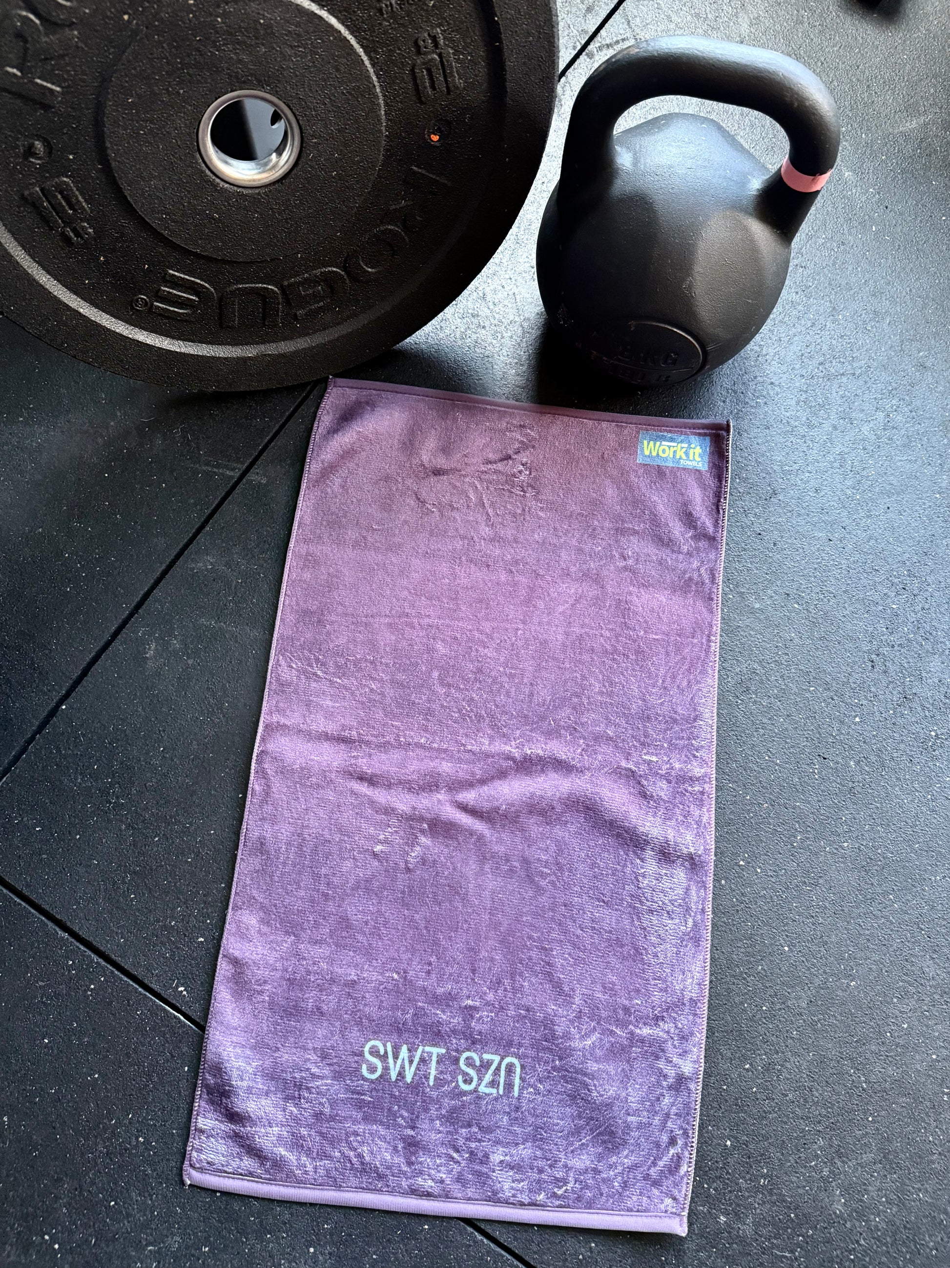 Purple towel with 'SWT SZN' branding on a black surface with gym equipment.